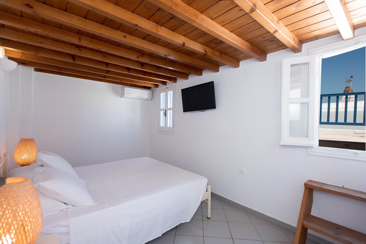 Un dormitorio con techo de madera, cama con ropa de cama blanca, televisor montado en la pared y ventana con vista a un balcón.