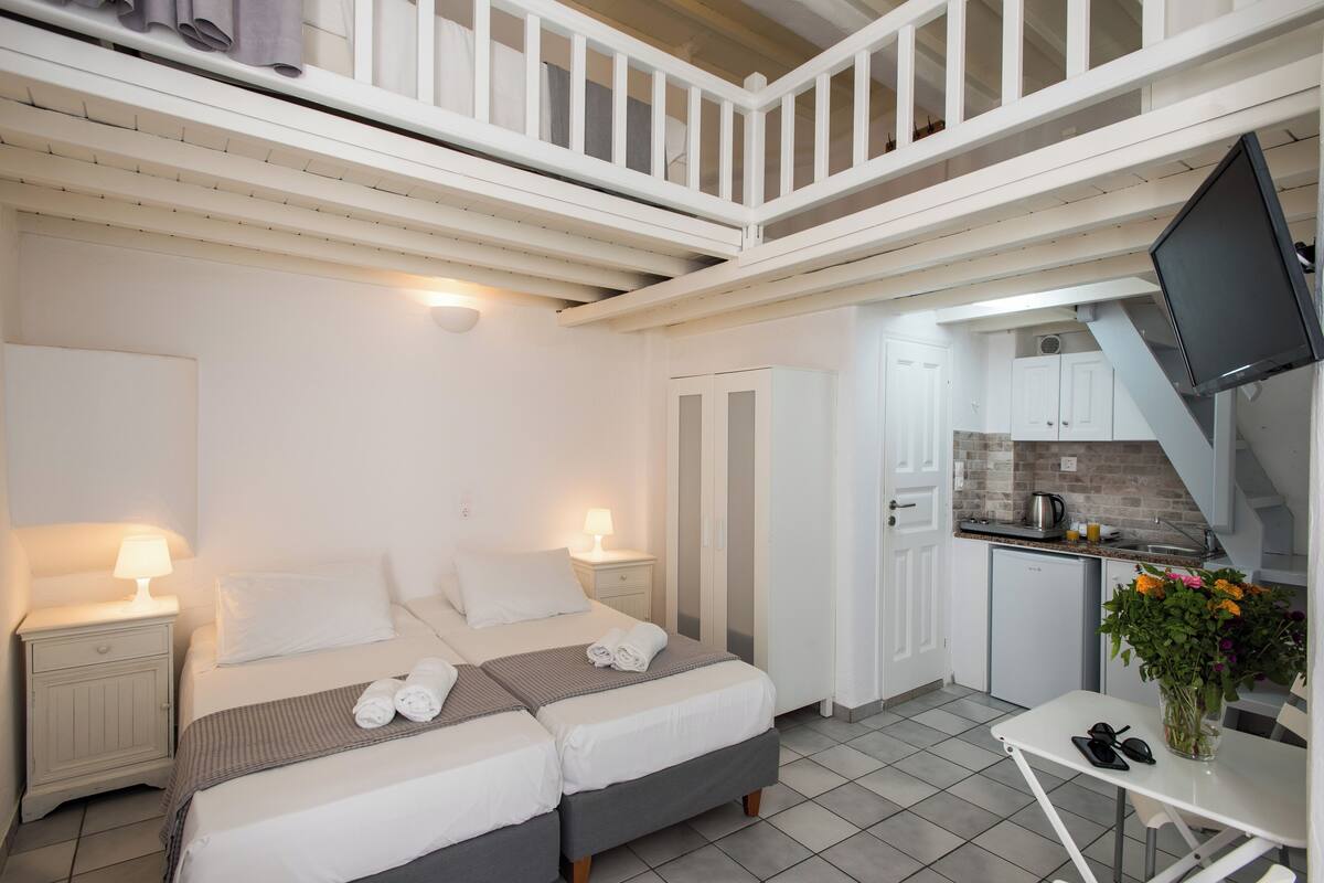 A bedroom with two beds, a small table with a vase of flowers, a kitchenette, and a balcony.
