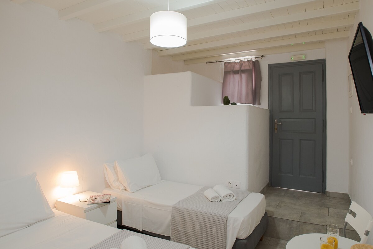 Habitación de hotel con dos camas, una mesita con vasos de jugo de naranja, una silla blanca, un televisor en la pared y una puerta con cortina.