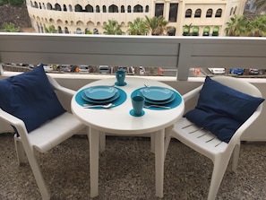 Varanda com mesa posta para dois, com cadeiras brancas e almofadas azuis.