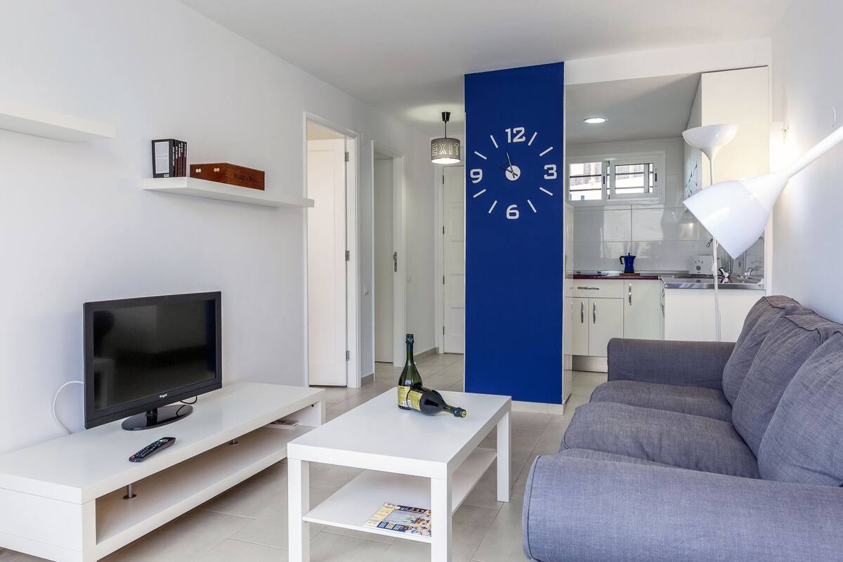 Ein modernes Wohnzimmer mit einem Flachbildfernseher, einer Fernbedienung, einem kleinen Couchtisch mit einer Flasche, einem grauen Sofa und einem weißen Couchtisch. Eine blaue Wanduhr ist sichtbar, und im Hintergrund ist eine Küchenzeile zu sehen.