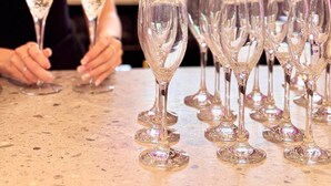 Una zona de bar con varias copas de champán dispuestas sobre una barra, una mujer sosteniendo una copa y un cartel que dice 'BLOOMS BAR'.