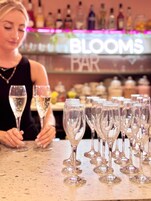 Una zona de bar con varias copas de champán dispuestas sobre una barra, una mujer sosteniendo una copa y un cartel que dice 'BLOOMS BAR'.