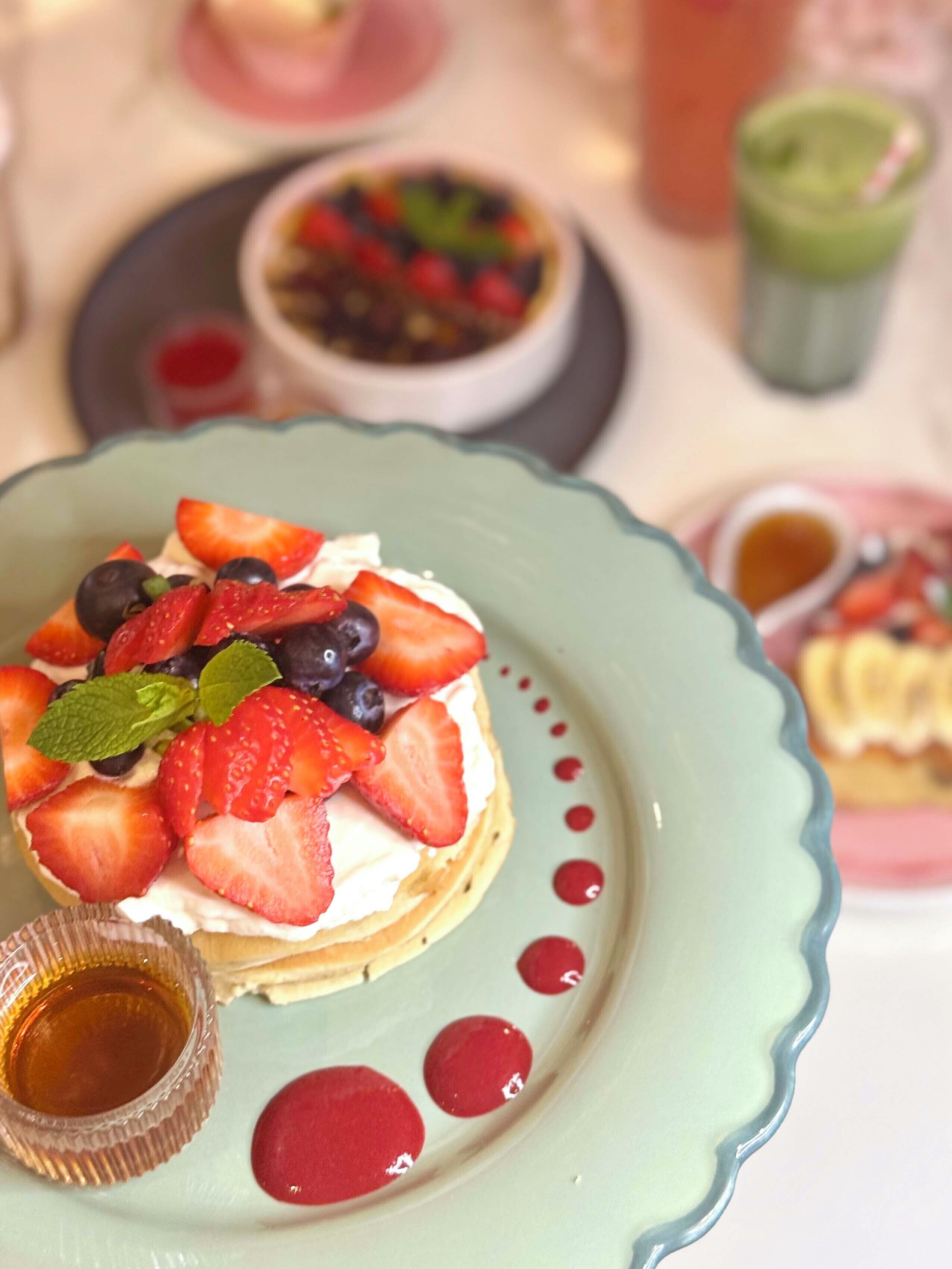 A dessert plate with strawberries, blueberries, and mint on a light blue plate, accompanied by a small jar of syrup and red dots.