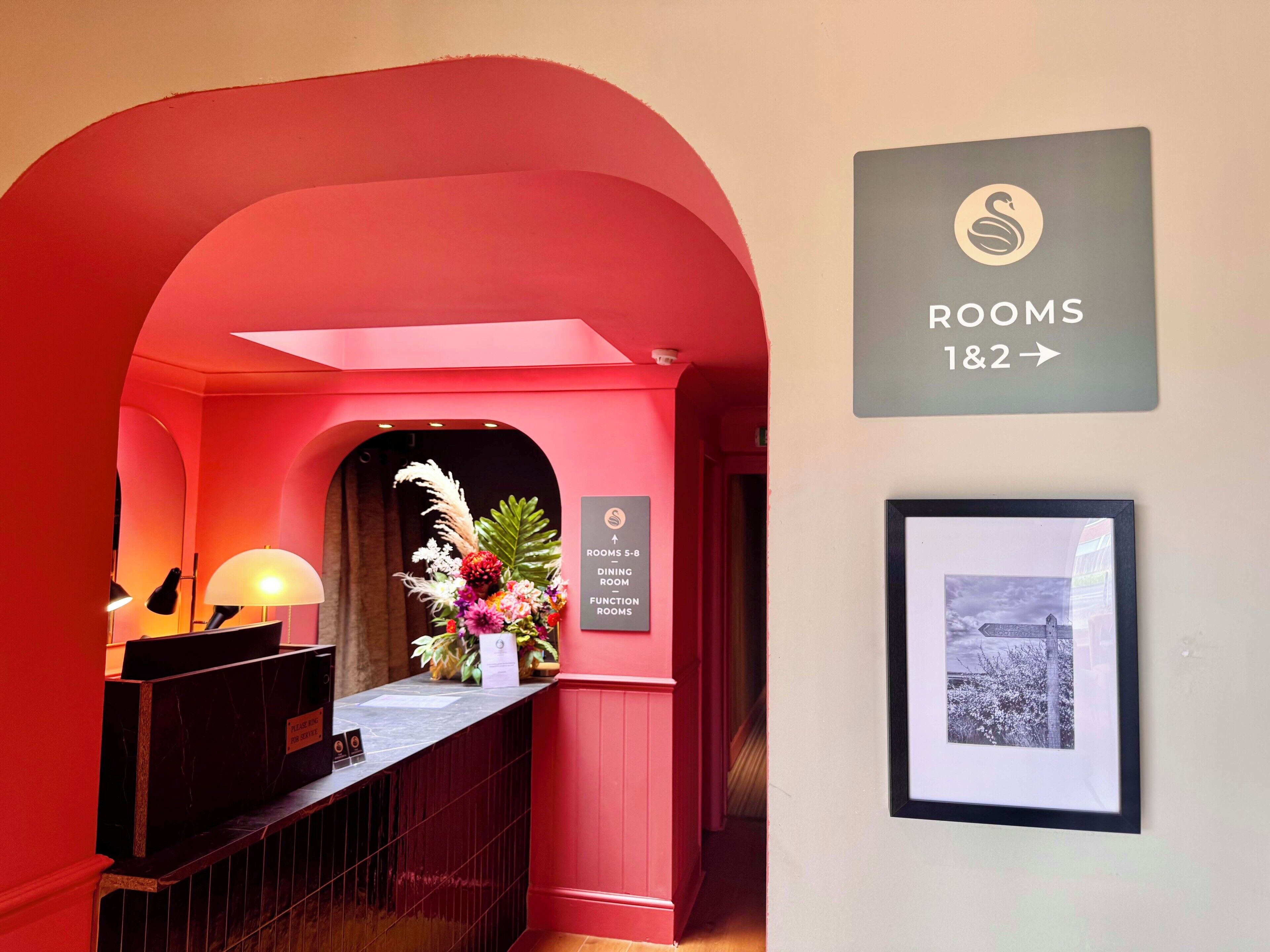 A reception area with a red archway, a desk with a lamp, a framed picture, and a sign indicating 'ROOMS 1&2' and 'DINING ROOM FUNCTIONAL ROOMS.'