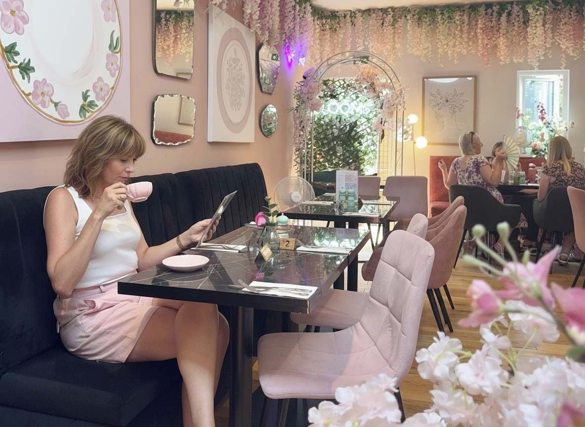 A woman sitting on a couch at a table with a tablet and a cup, in a room with floral decorations and a Christmas tree.