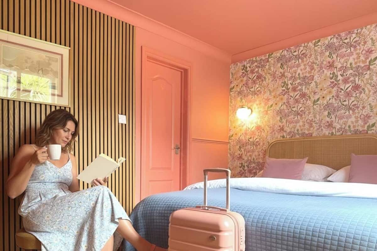 A room with a bed, a wall with floral wallpaper, a wooden panel wall, a door, and a suitcase.
