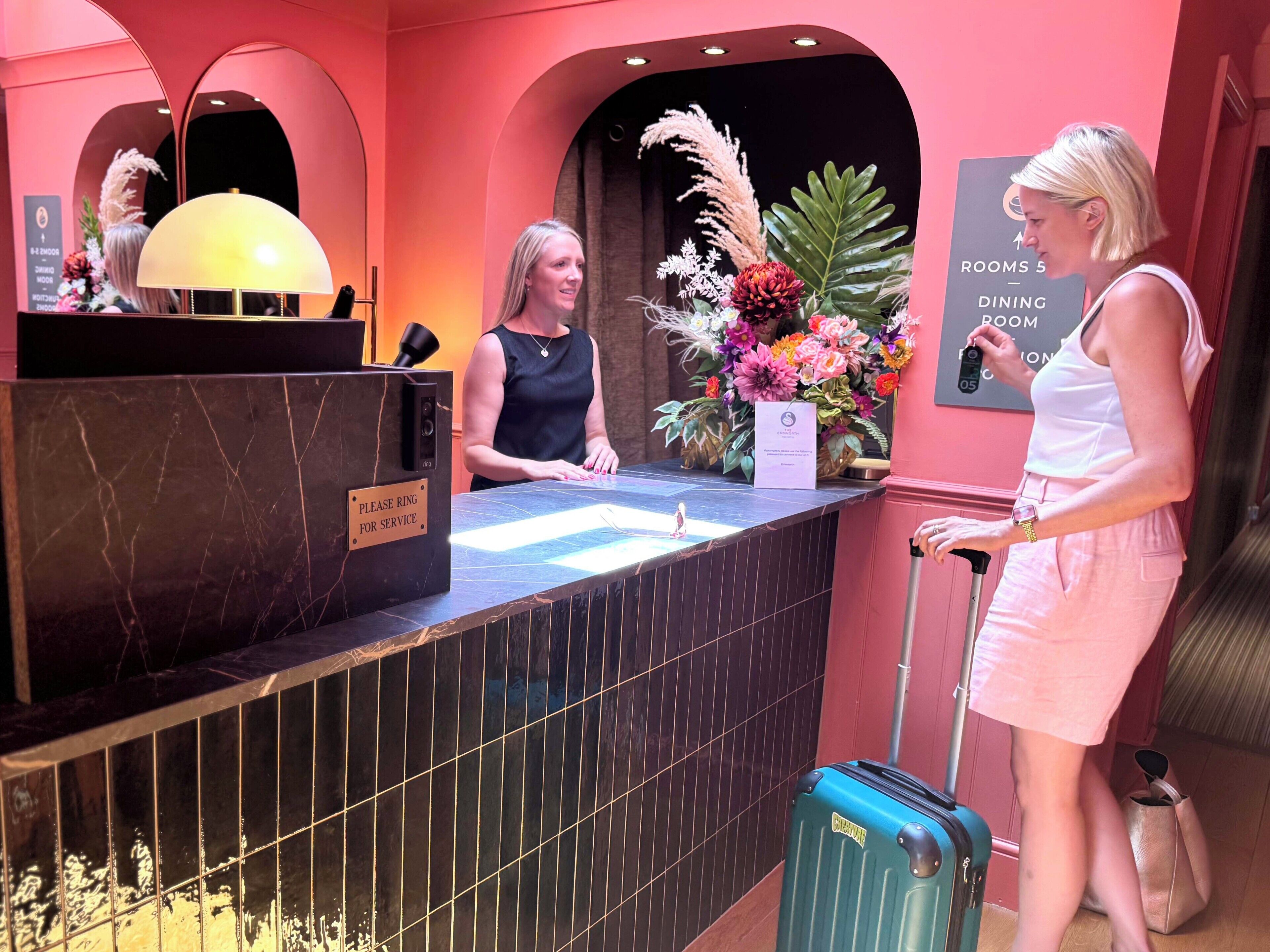 Un comptoir de réception d’hôtel avec une pancarte indiquant « Veuillez sonner pour être servi », un arrangement floral et une autre pancarte indiquant « Chambres 5 – Restaurant ».