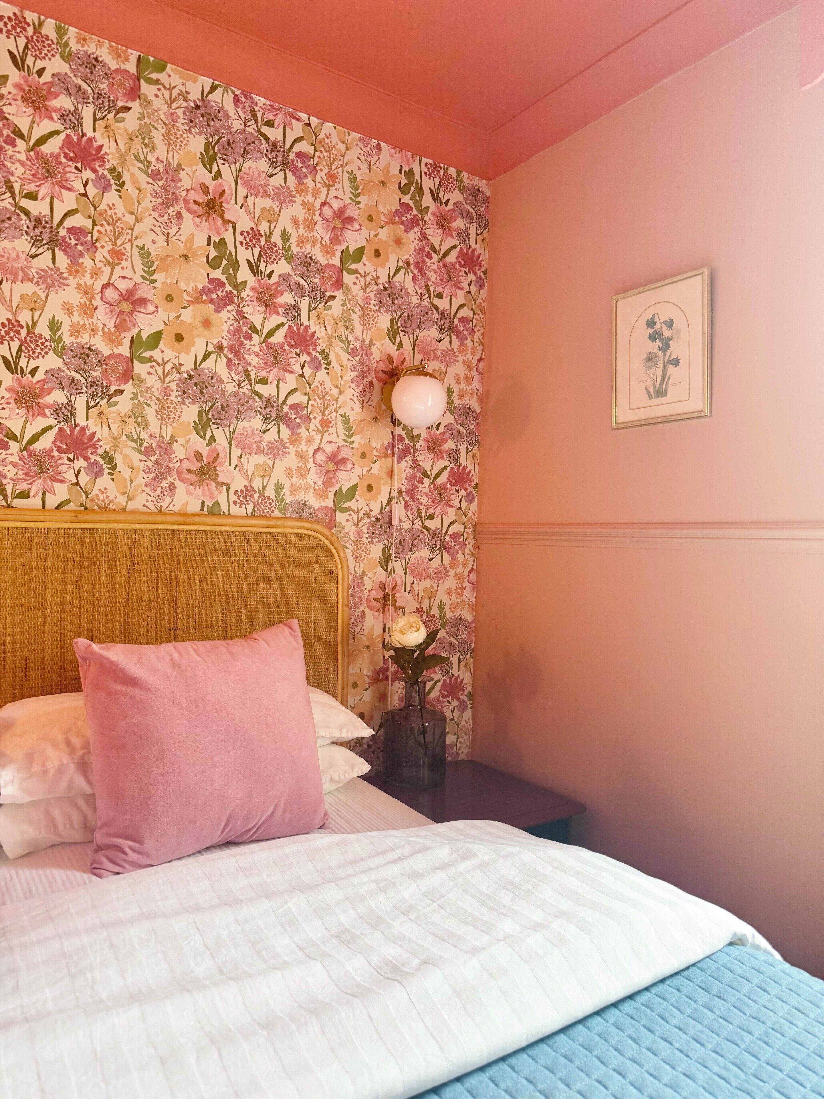 A bedroom with a floral wallpaper, a bed with pink and white bedding, a bedside lamp, and a framed picture on the wall.