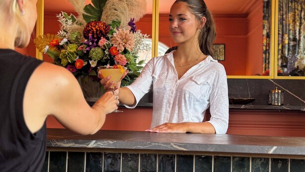 Una zona de bar con mostrador, un arreglo floral y un espejo que refleja la habitación.