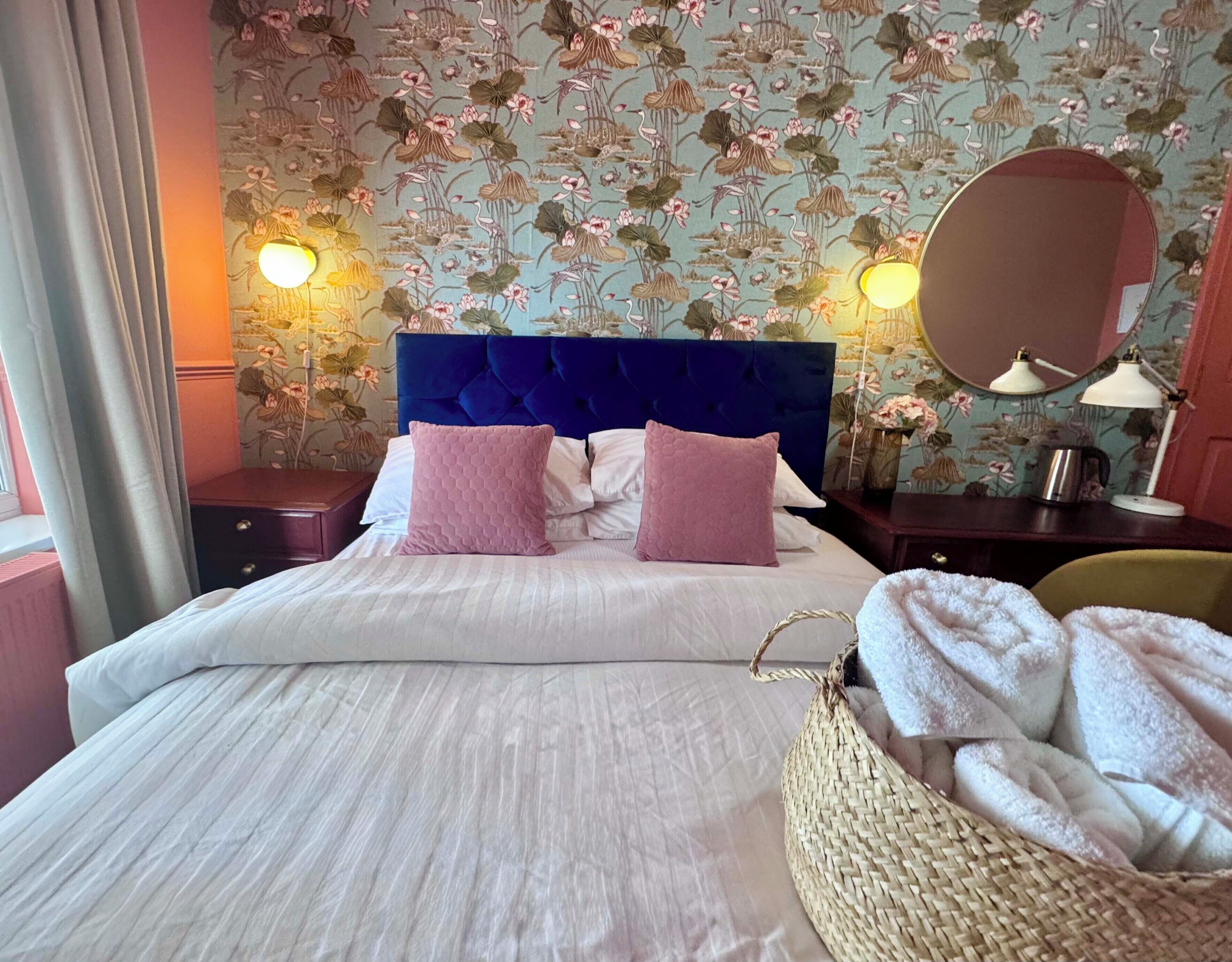 Un letto rifatto con cura, con una testiera blu, due cuscini rosa, un piumone bianco, un cesto in vimini con asciugamani, uno specchio rotondo e due lampade da comodino.
