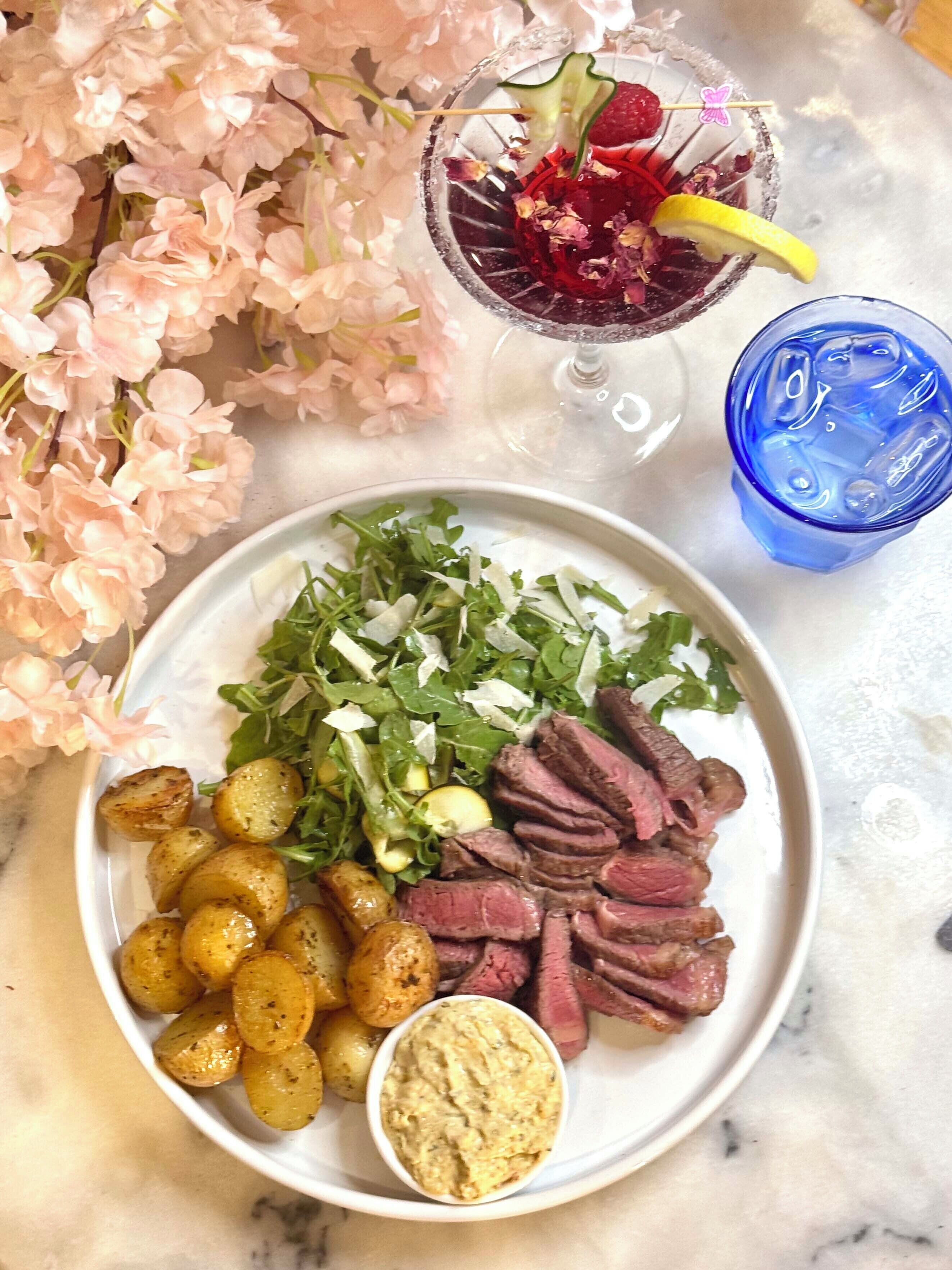 A plate with sliced meat, greens, and potatoes, a glass with a drink, and a blue glass with ice.