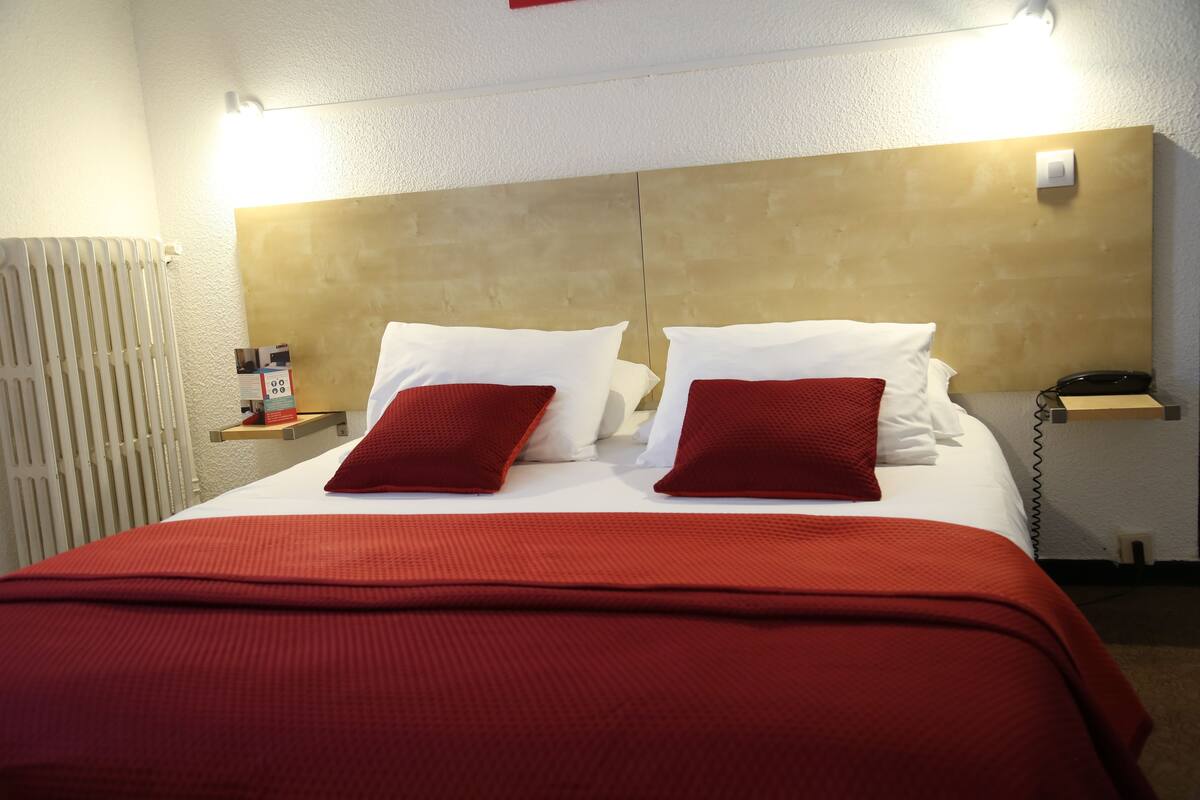 A hotel room with a large bed, red bedding, a wooden headboard, a bedside table with a phone, and a nightstand with a lamp.