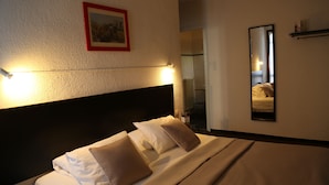 Una cama bien tendida con almohadas y cabecera, un cuadro enmarcado en la pared y un espejo que refleja parte de la habitación.
