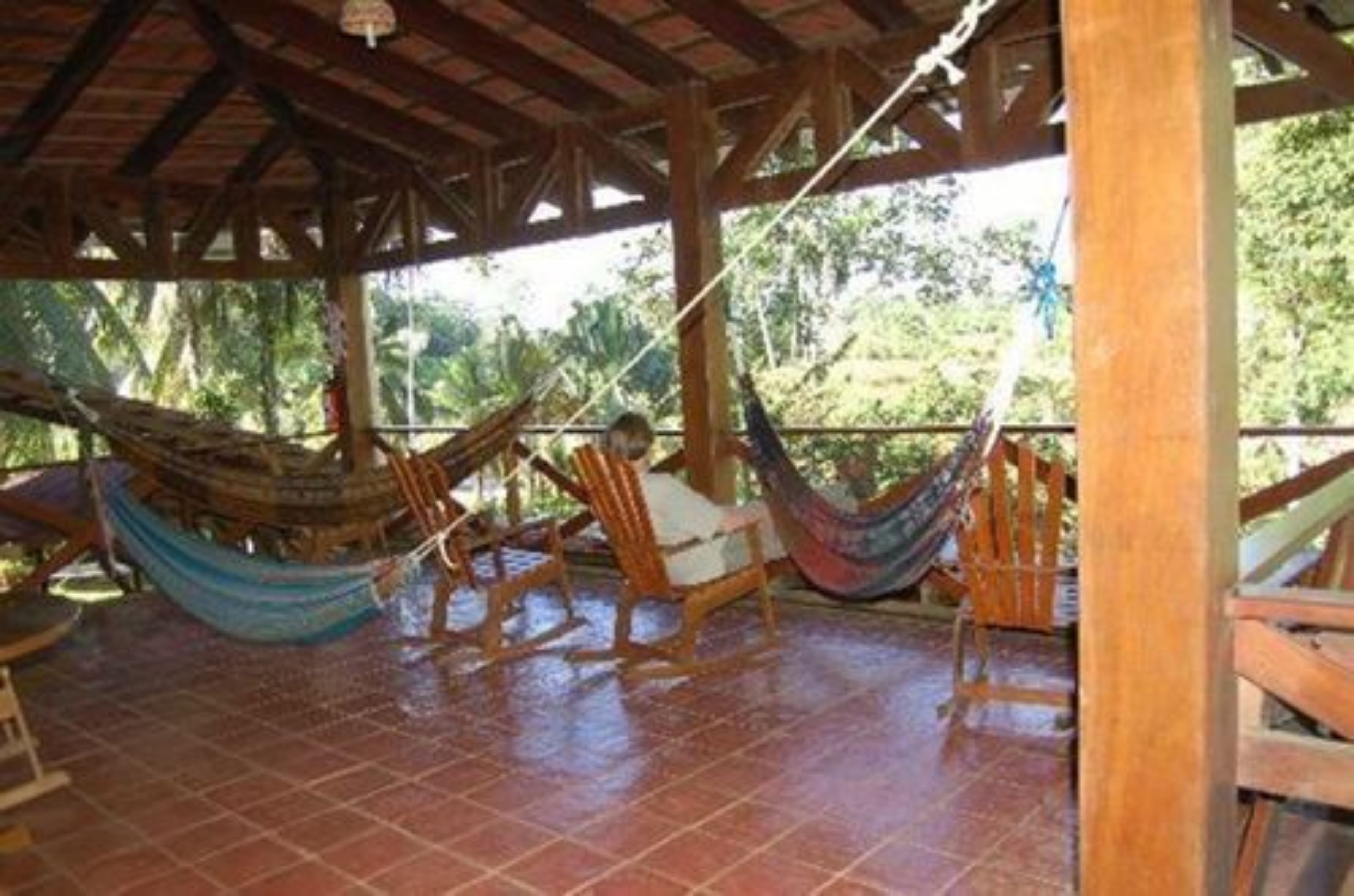 A covered outdoor area with wooden chairs and hammocks, surrounded by a tiled floor and wooden beams.