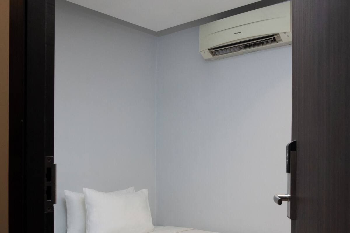 Ein kleines, ordentlich bezogenes Bett in einem Zimmer mit einer Holztür und einer Wand-Klimaanlage.