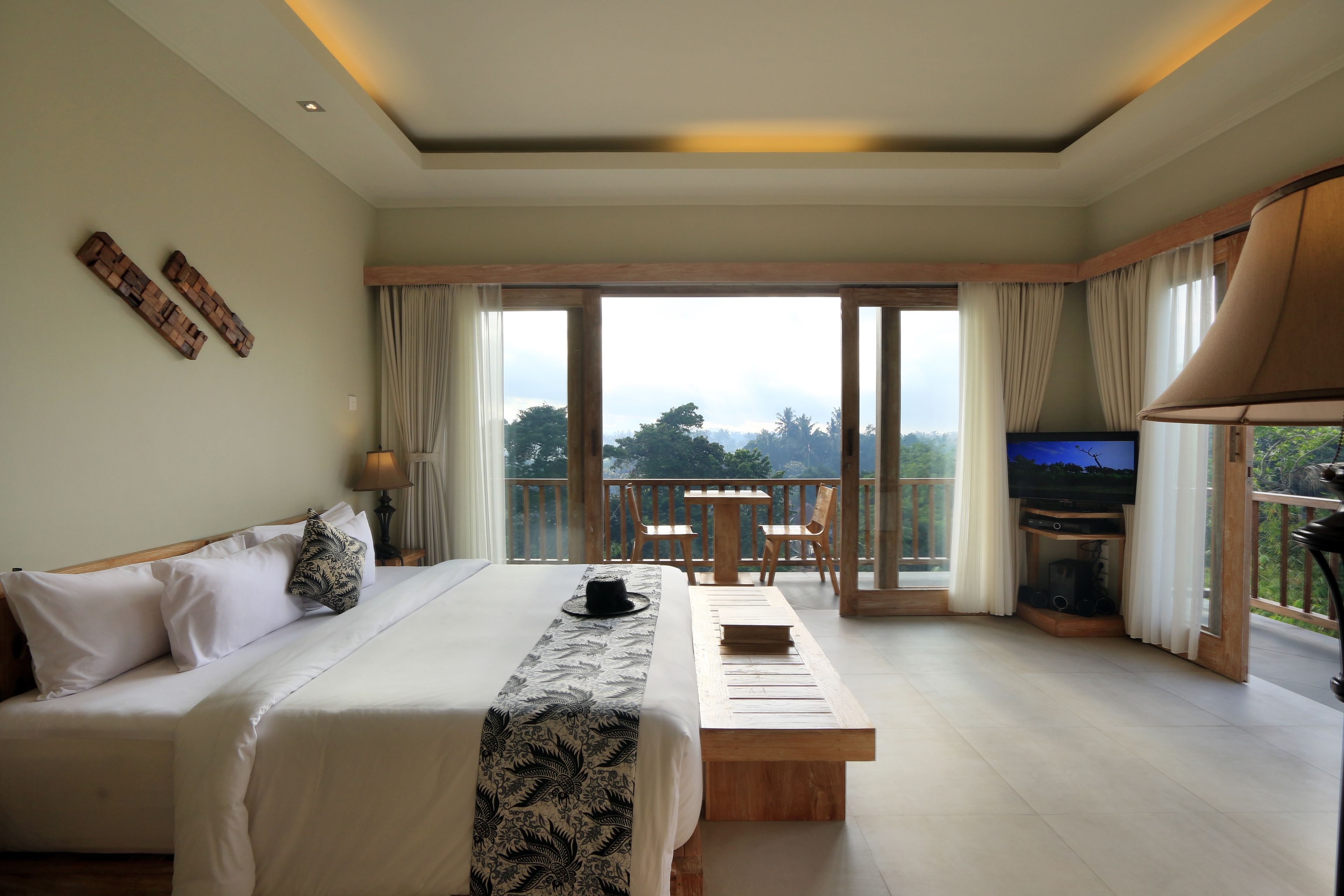 Ein Schlafzimmer mit einem großen Bett, einer hölzernen Bank, einem Fernseher und einem Balkon mit Blick auf Bäume.