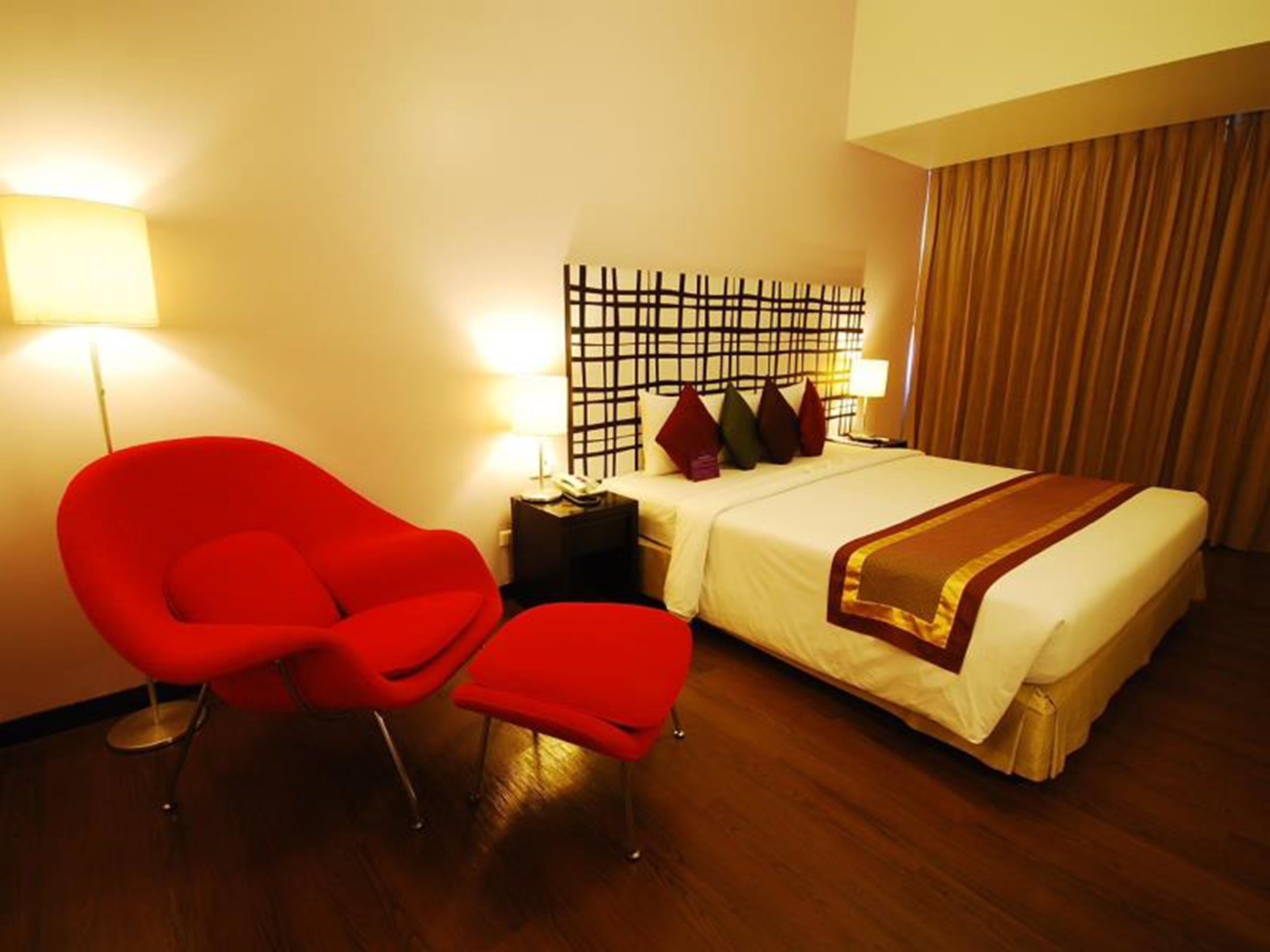 A hotel room with a bed, a red armchair, a nightstand, and a wall-mounted light fixture.
