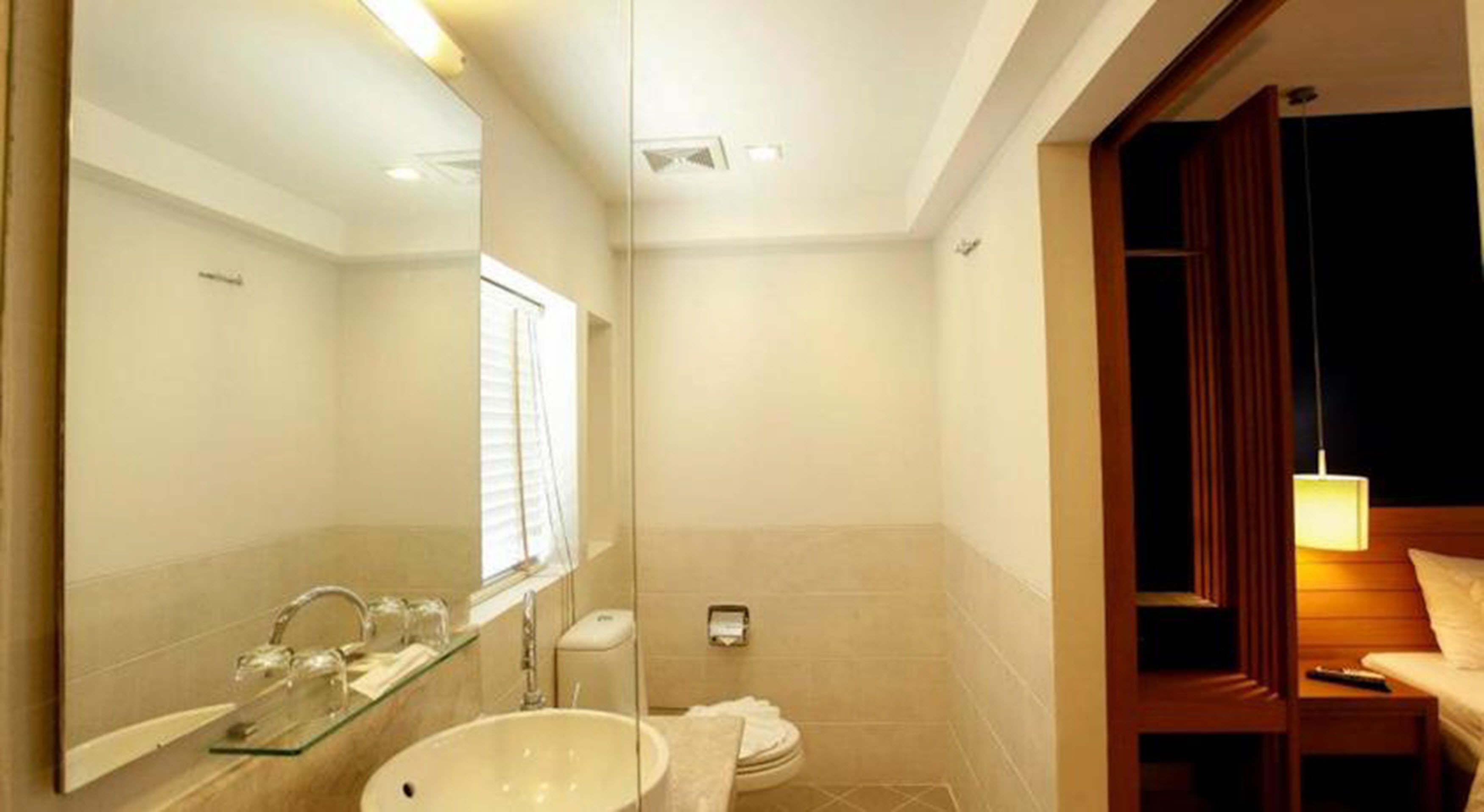 A modern bathroom with a large mirror, a sink, and a toilet. There is a window with blinds, a towel rack, and a wooden bench with a lamp.
