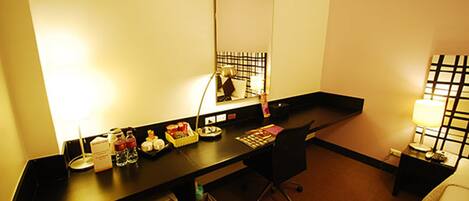 A hotel room with a desk, chair, bedside table, lamp, and a bed.