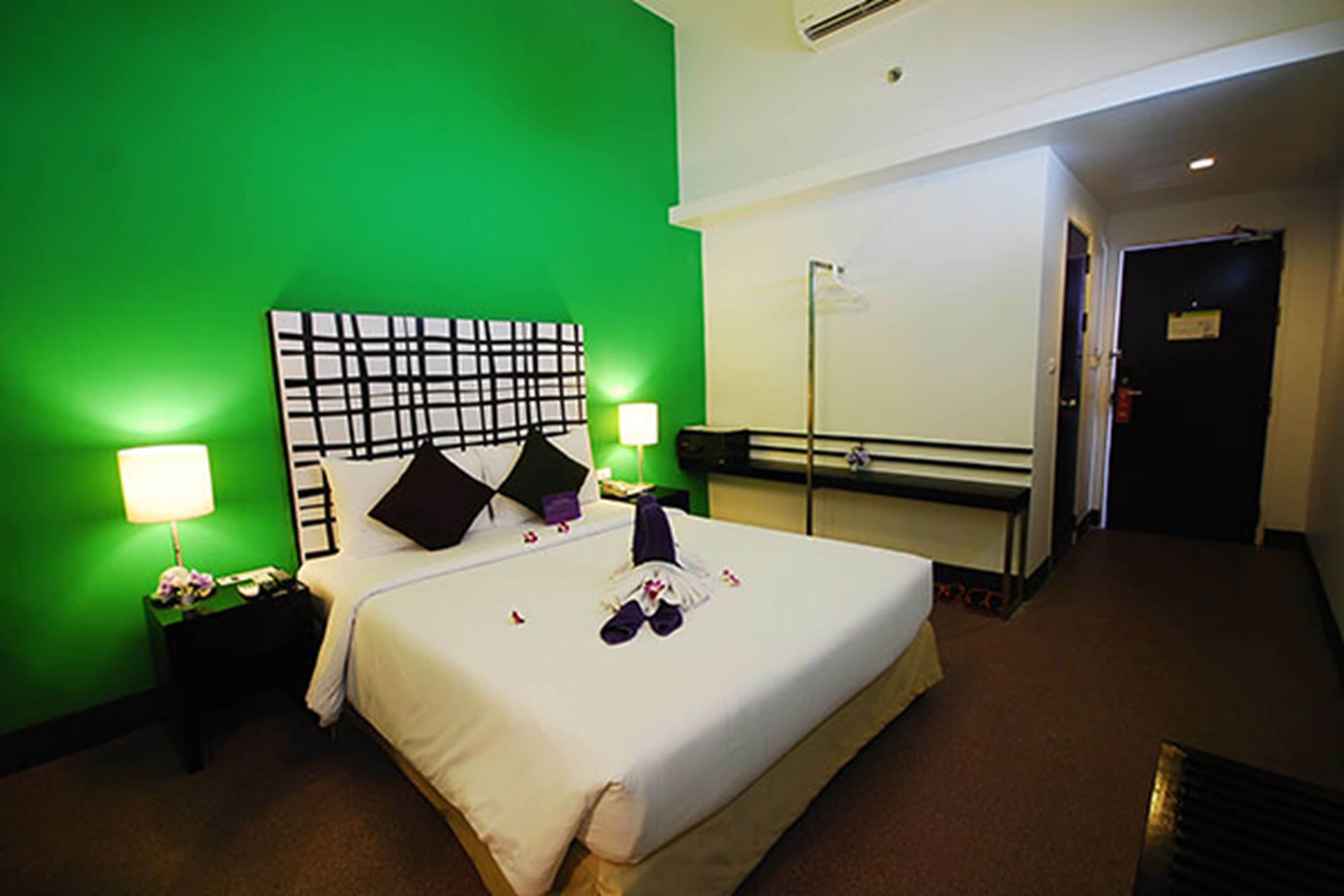 Habitación de hotel con pared verde, una cama grande con ropa de cama blanca, dos mesitas de noche con lámparas y un cabecero blanco y negro.