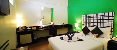A hotel room with a green accent wall, a bed with a heart-shaped towel arrangement, a desk with a mirror, and a TV on the wall.
