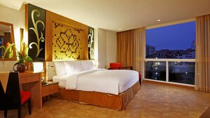 Hotelkamer met een groot bed, een bureau met stoel, een spiegel en uitzicht op de stad in de schemering.