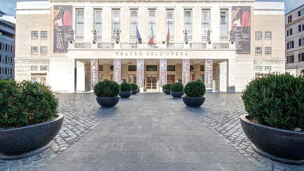 A grand entrance to Teatro dell'Opera with a symmetrical facade, large potted plants, and flags.