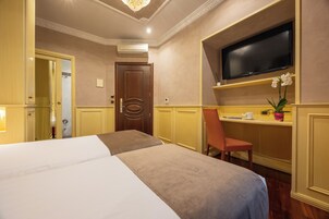 Una camera d'albergo con un letto, una televisione montata a parete, una scrivania con una sedia e una pianta in vaso.