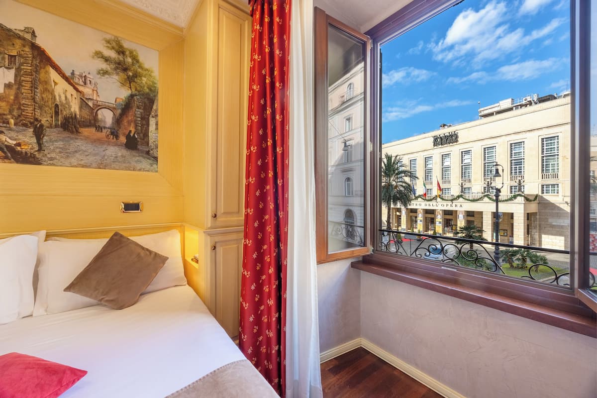 Habitación de hotel con cama, un cuadro en la pared, ventana con vista a un edificio y una cortina roja.