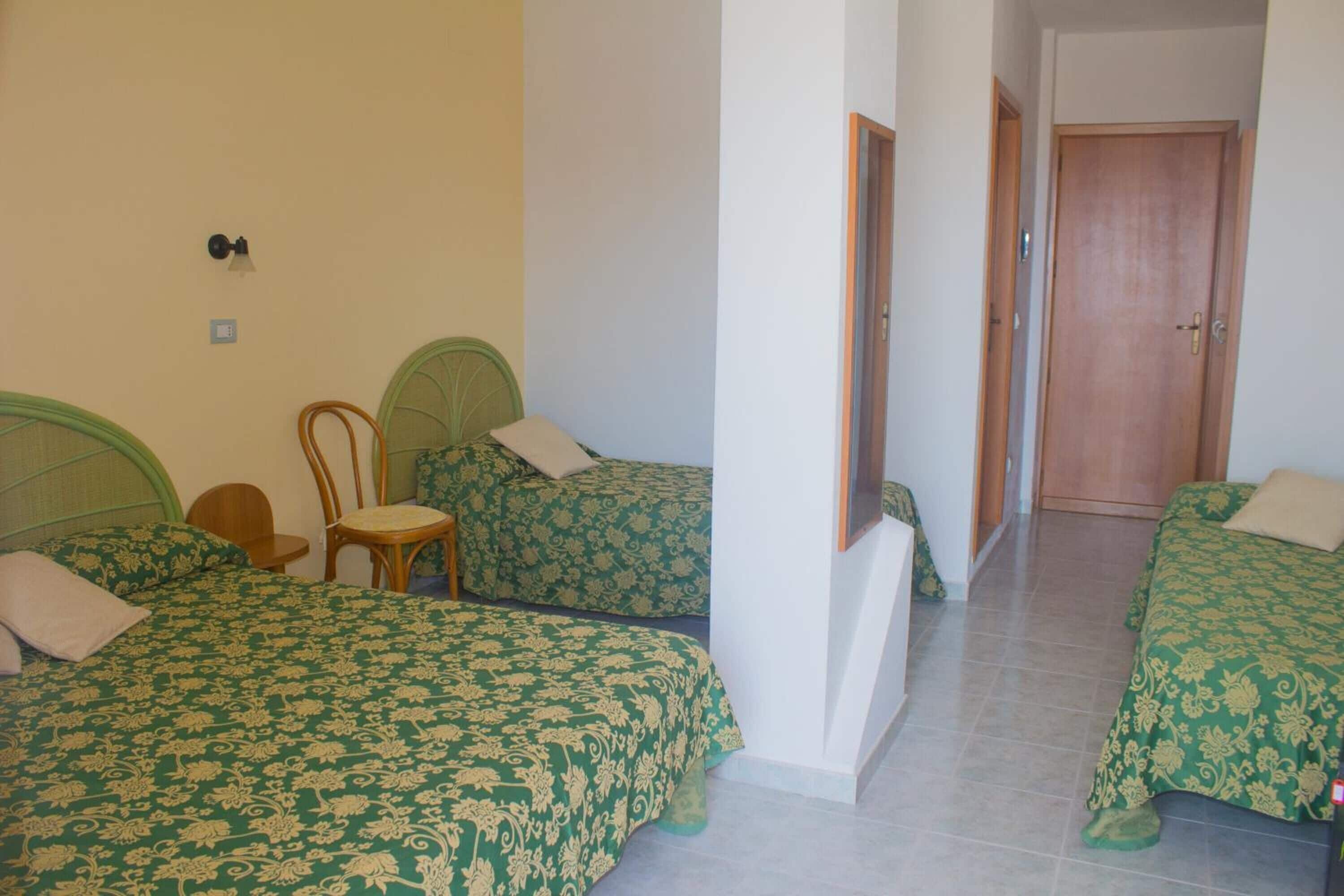 A room with two beds, each with green patterned bedding, a wooden chair, and a small table.