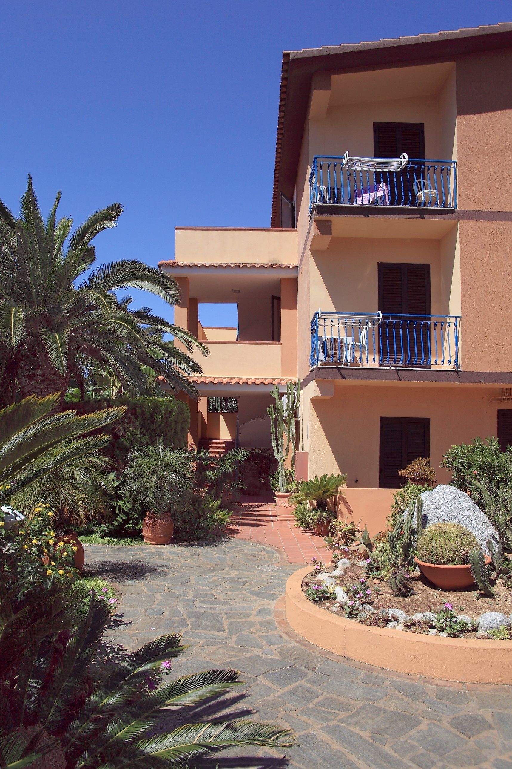 A multi-story building with a central courtyard, featuring a stone pathway, various potted plants, and a blue balcony railing.