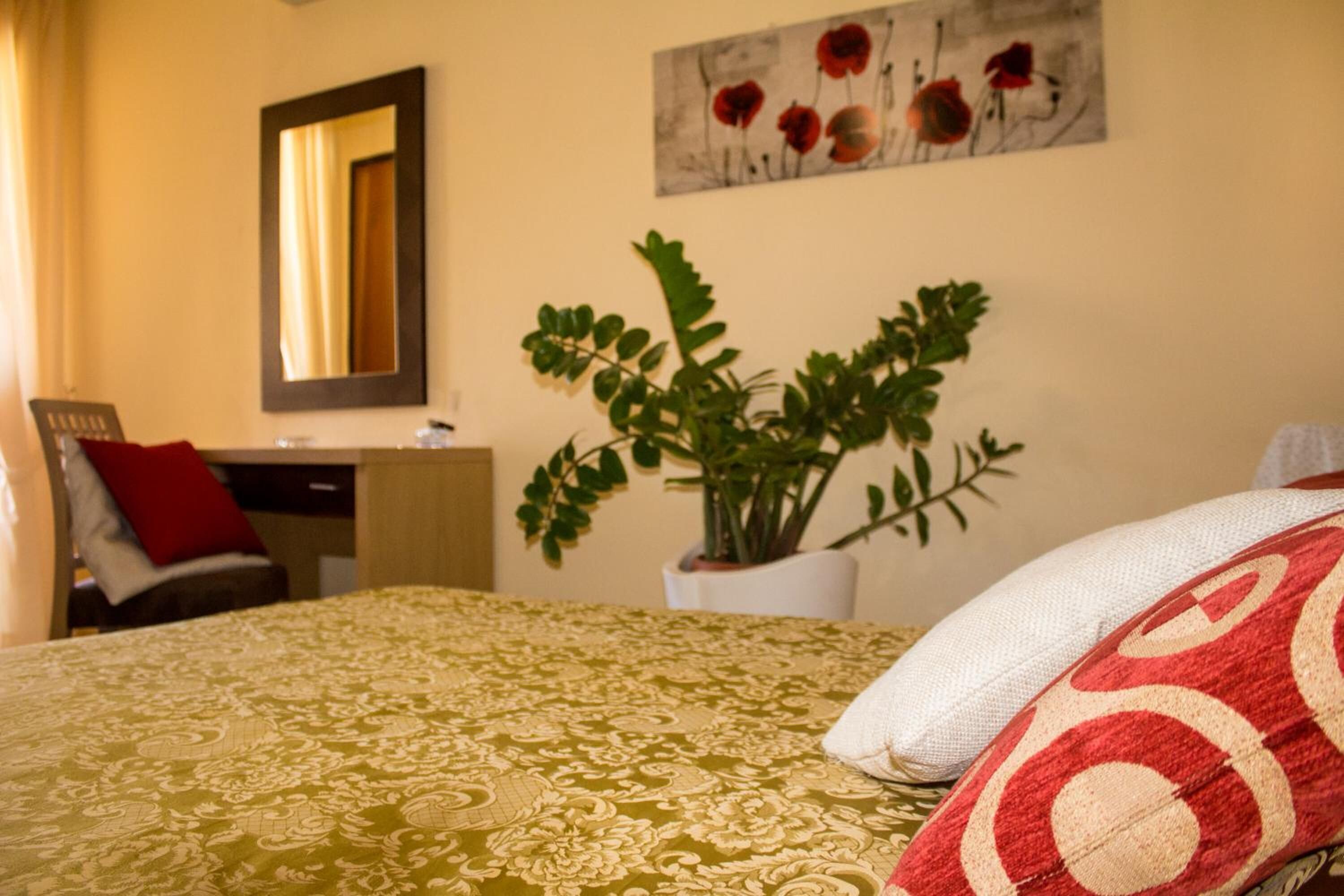 Una habitación de hotel con cama, escritorio, silla, espejo, planta y un cuadro en la pared.