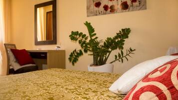 Una habitación de hotel con cama, escritorio, silla, espejo, planta y un cuadro en la pared.
