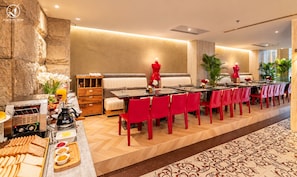 A hotel dining area with a long table, red chairs, a buffet with bread and spreads, and a coffee machine.