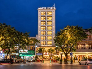 Un hôtel reconnaissable à son enseigne bien visible, avec de nombreuses fenêtres et une vue sur la rue, où l’on aperçoit des voitures garées et des arbres.