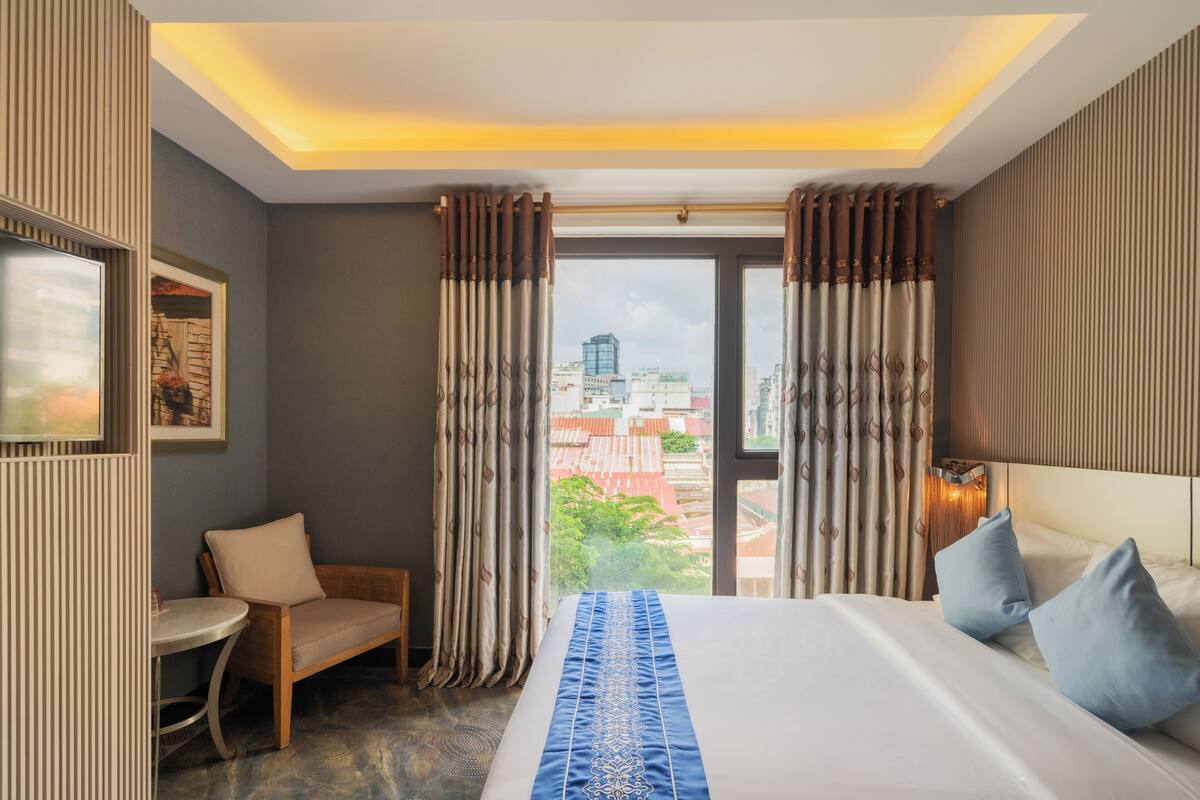Ein Hotelzimmer mit Bett, Sessel, kleinem Tisch und Blick auf die Stadt durch das Fenster.