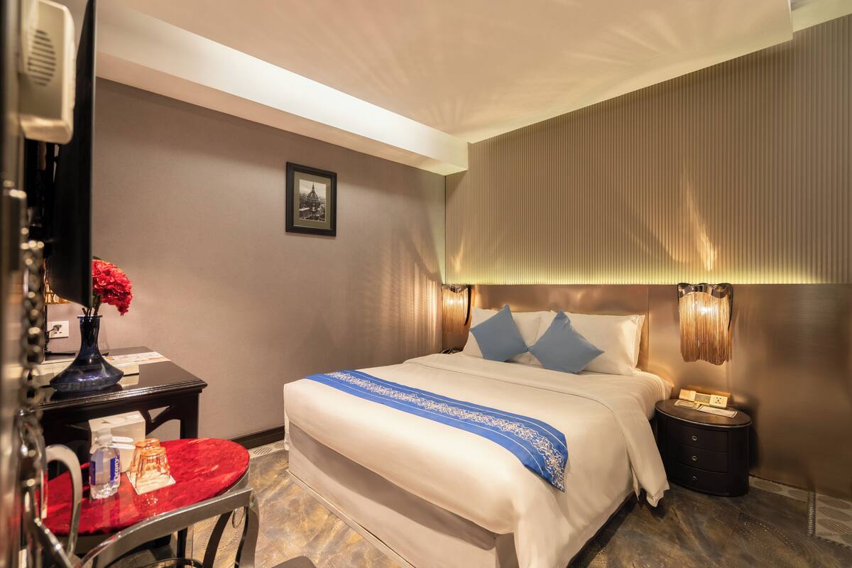 Ein Hotelzimmer mit einem ordentlich bezogenem Bett, einem Nachttisch mit einer Lampe, einem Sessel mit rotem Kissen und einem Bild an der Wand.