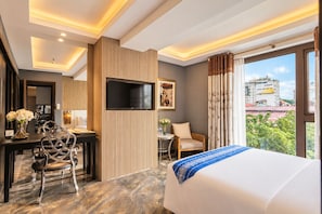 A hotel room with a bed, desk, chairs, a TV, and a view of the city.