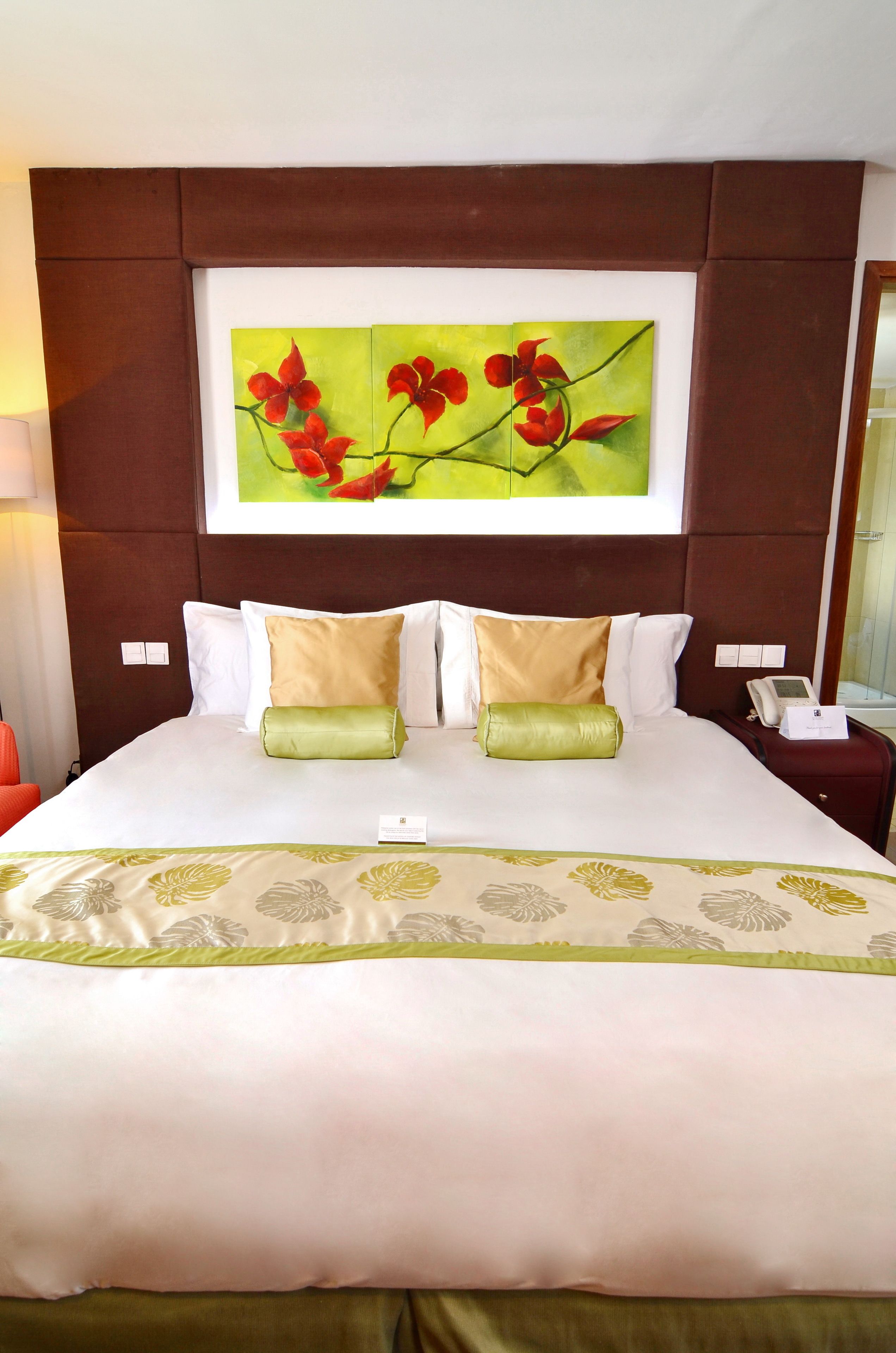 A neatly made bed with a floral bedspread, a headboard with a framed painting, and a bedside table with a phone and a lamp.