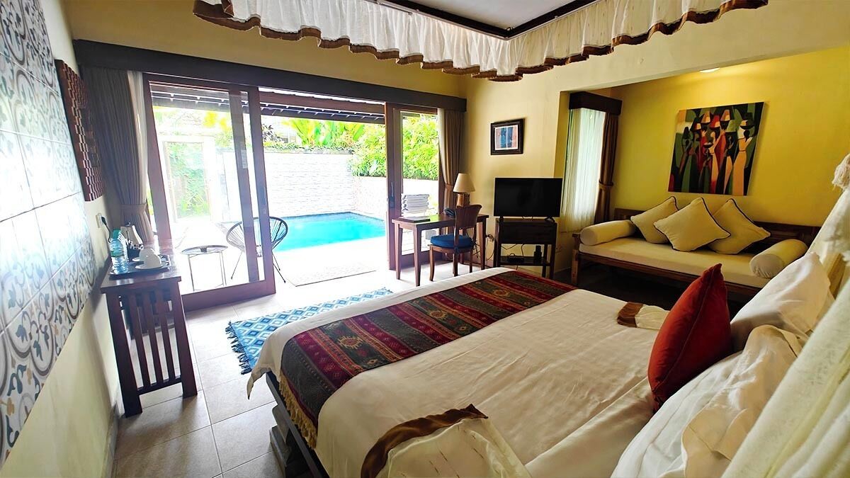A hotel room with a large bed, a TV, a sofa, and a desk. There is a sliding glass door leading to a pool area.