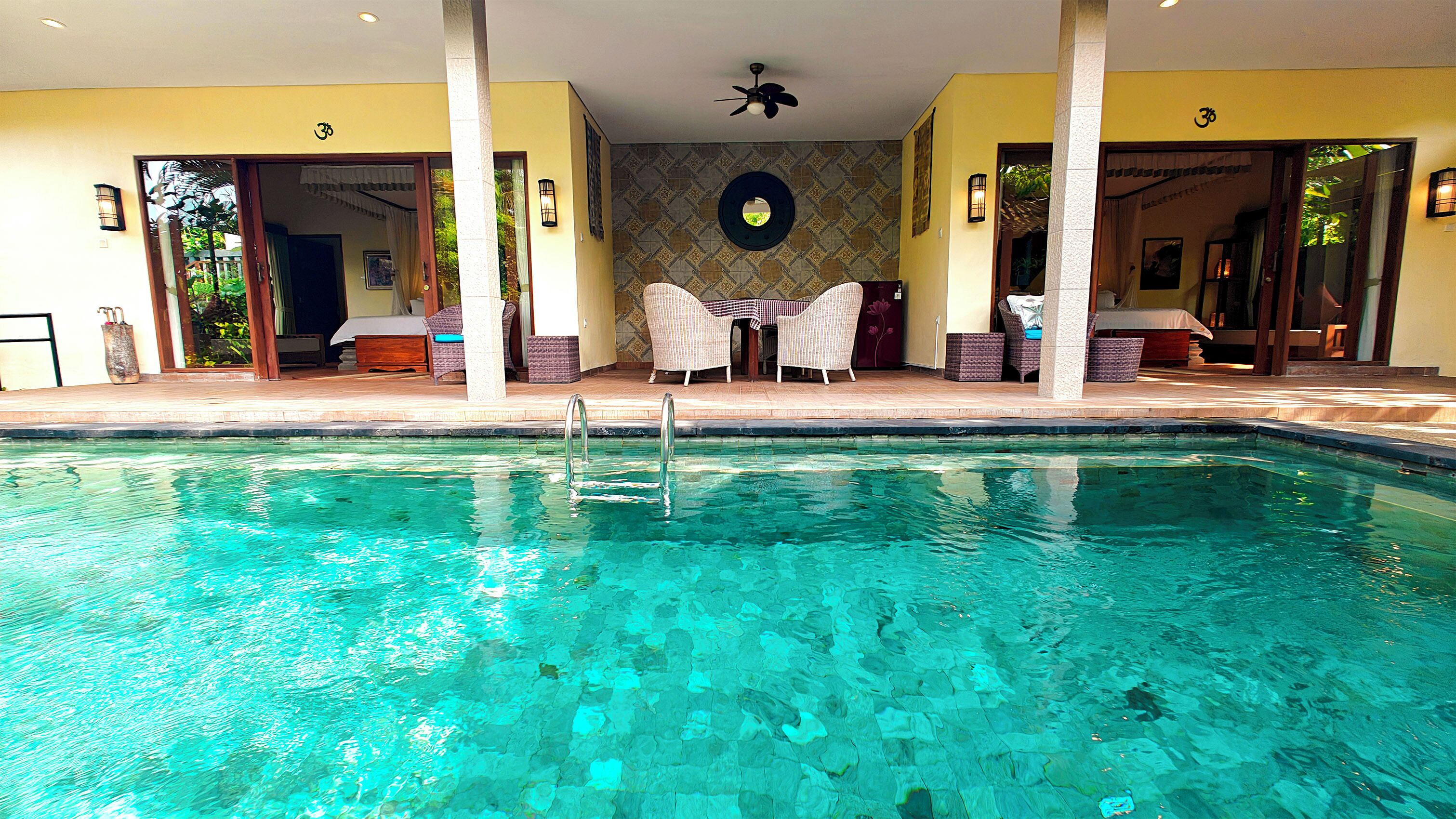 Une piscine aux eaux cristallines, dotée d’un parasol et de chaises longues, entourée d’un espace abrité avec un mur décoré et un ventilateur de plafond.