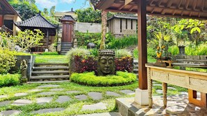 Un patio tradicional con escalones de piedra que conducen a una puerta de madera que dice “Recepción y Caja”, rodeado de abundante follaje y una estatua de piedra.