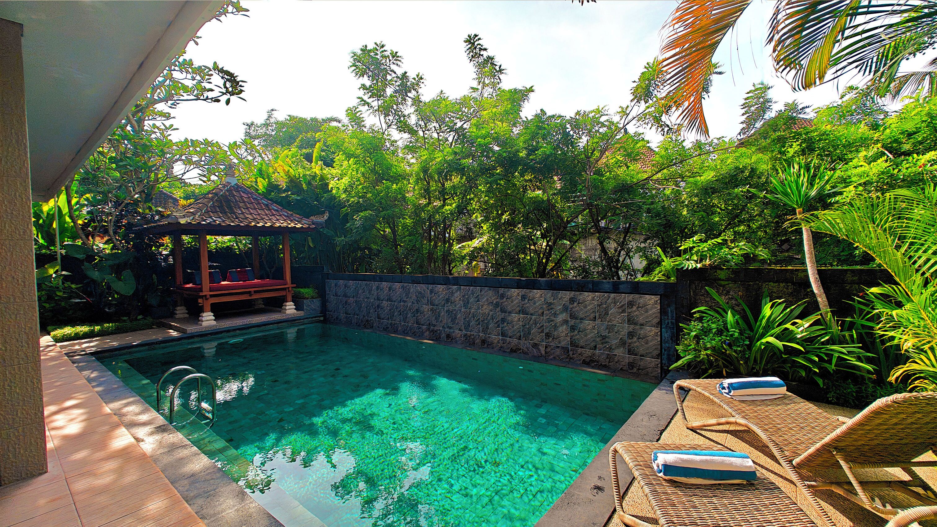 A swimming pool with clear water, surrounded by lush greenery and a wooden gazebo.
