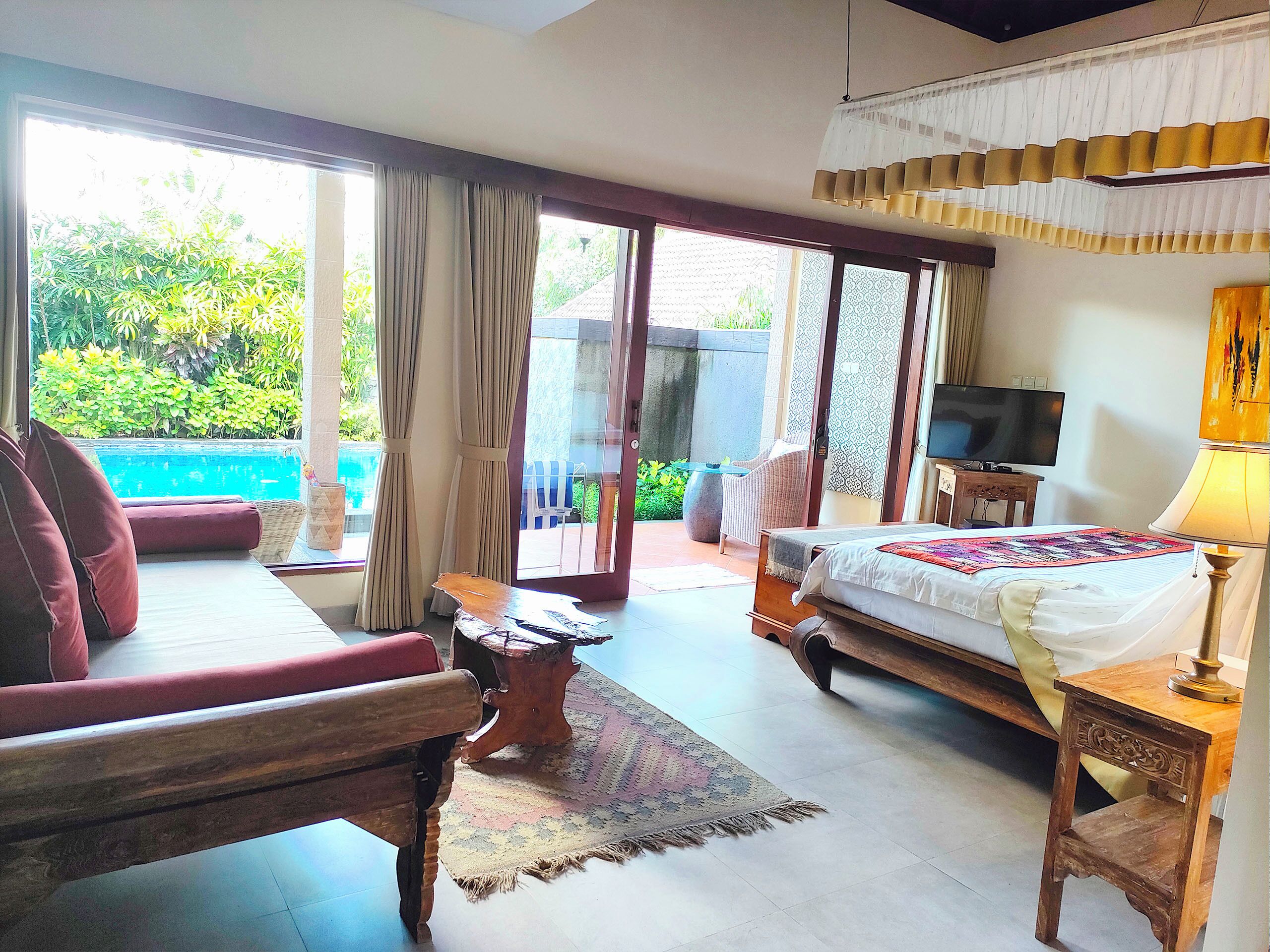 A bedroom with a bed, a TV, a wooden chair, a small table, and a view of a swimming pool.