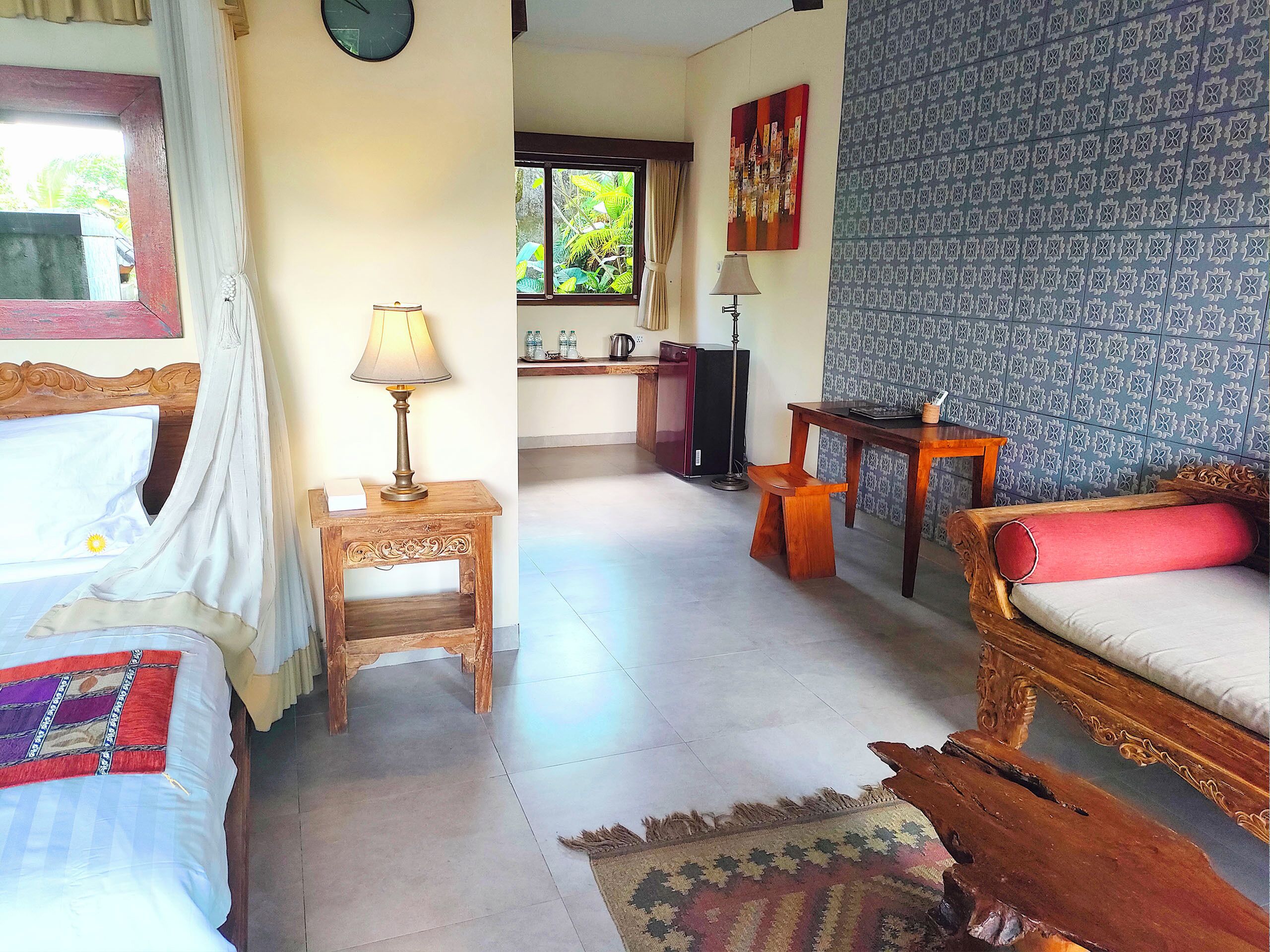 A hotel room with a bed, a small table with a lamp, a wooden bench, a sofa, a patterned wall, and a window with curtains.