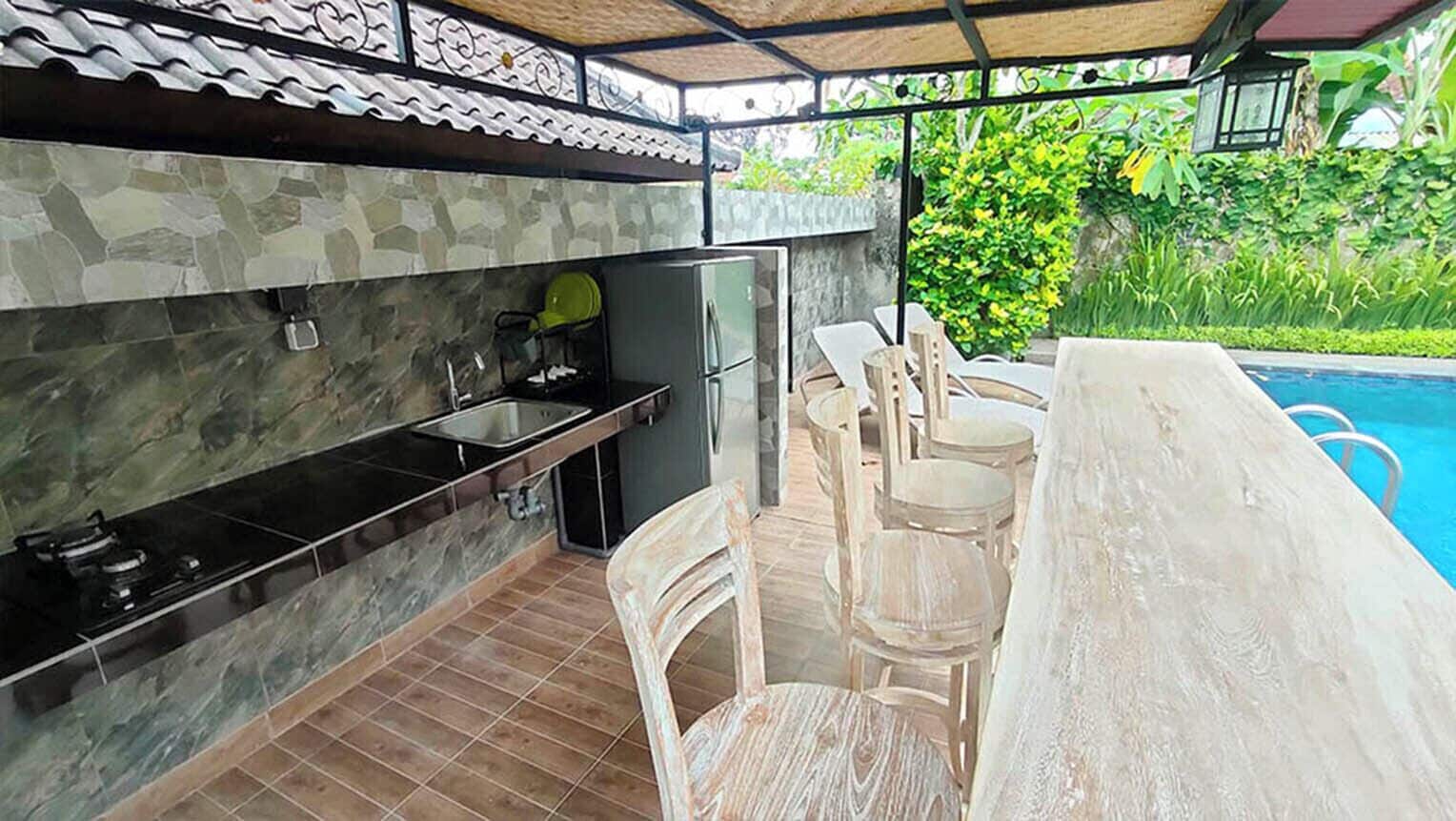 Cocina exterior con protector mural de piedra, pileta y refrigerador. Hay sillas y una mesa de comedor cerca de una piscina.