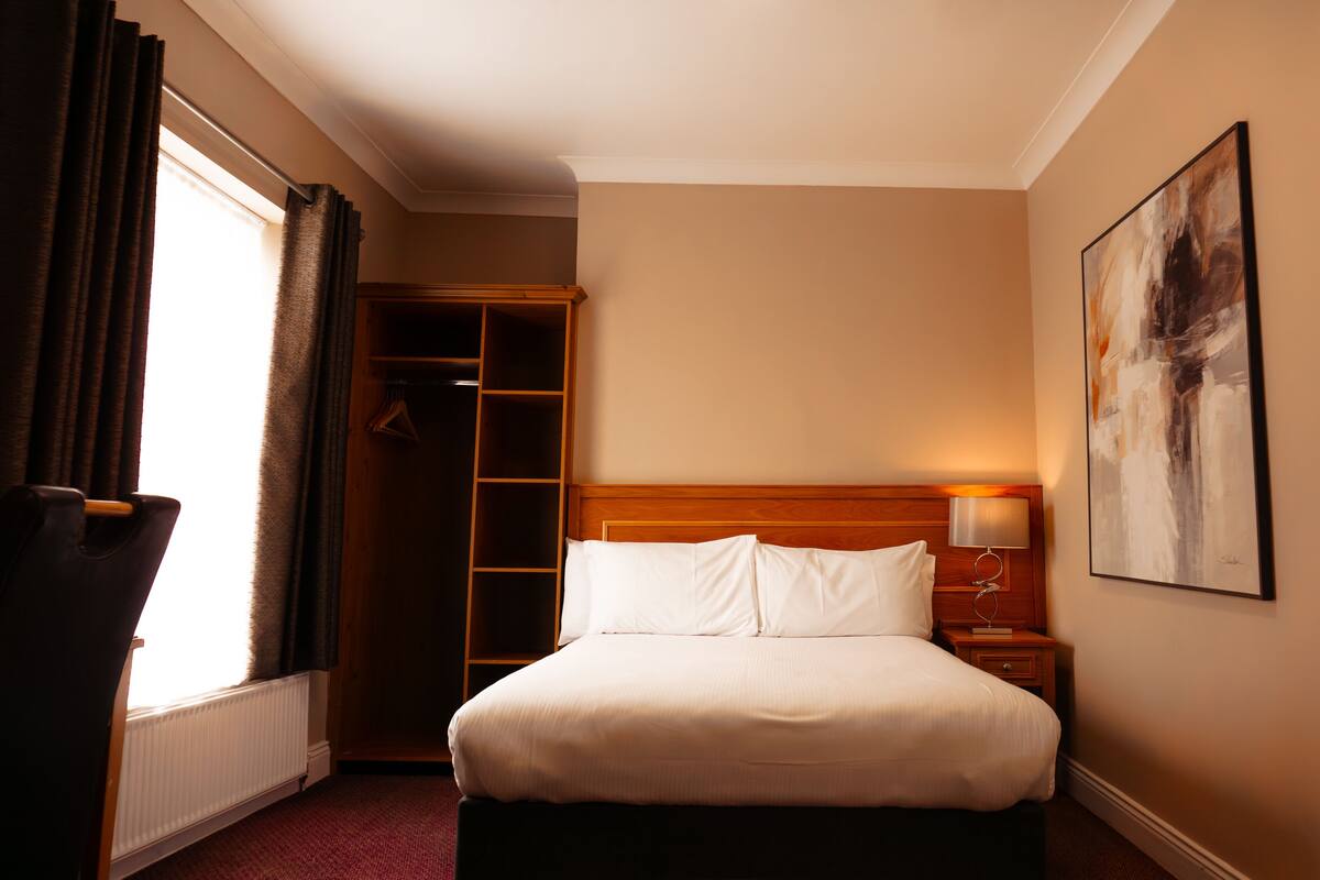 A hotel room with a large bed, a wooden headboard, a nightstand, a lamp, a closet, and a painting on the wall.