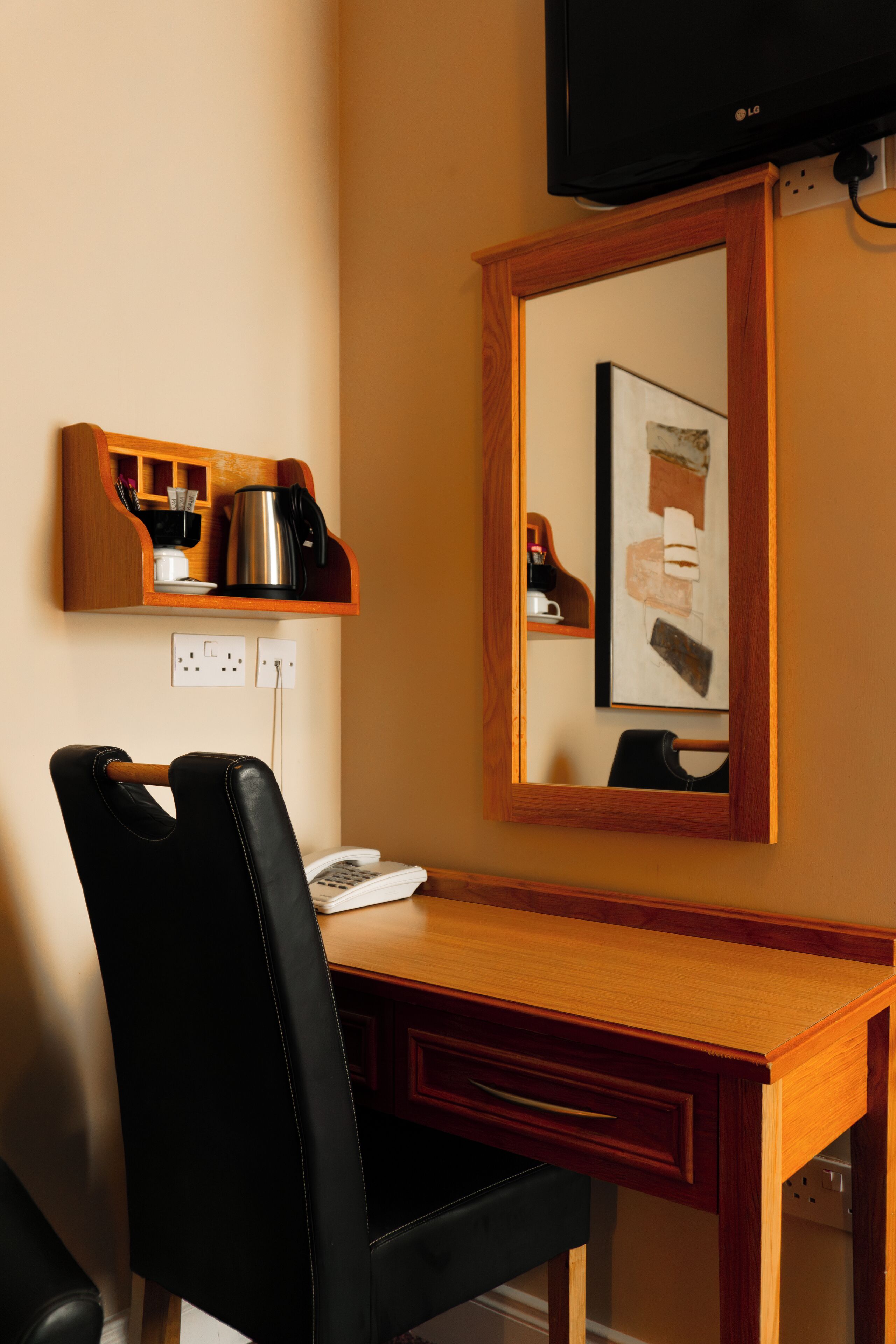 A hotel room with a wooden desk, a black leather chair, a coffee maker, a telephone, and a framed artwork on the wall.