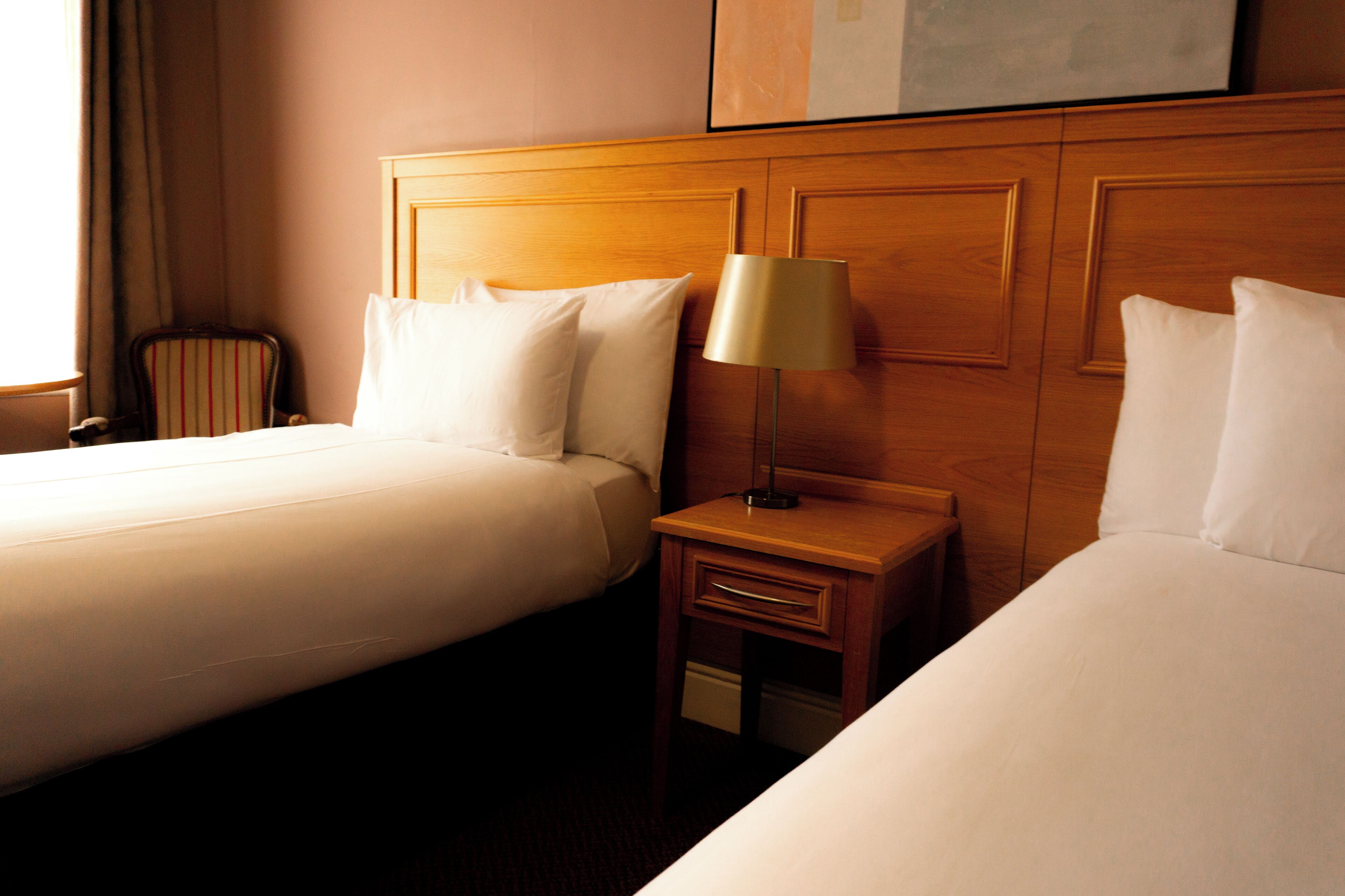 Habitació d'hotel amb dos llits, un capçal de fusta, una taula de nit amb una làmpada i una cadira.